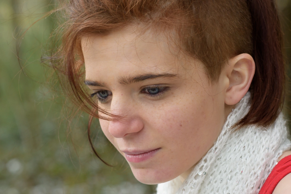 Portrait rapproché d’une femme en extérieur, regard vers le bas, cheveux légèrement au vent, lumière naturelle douce et arrière-plan flou